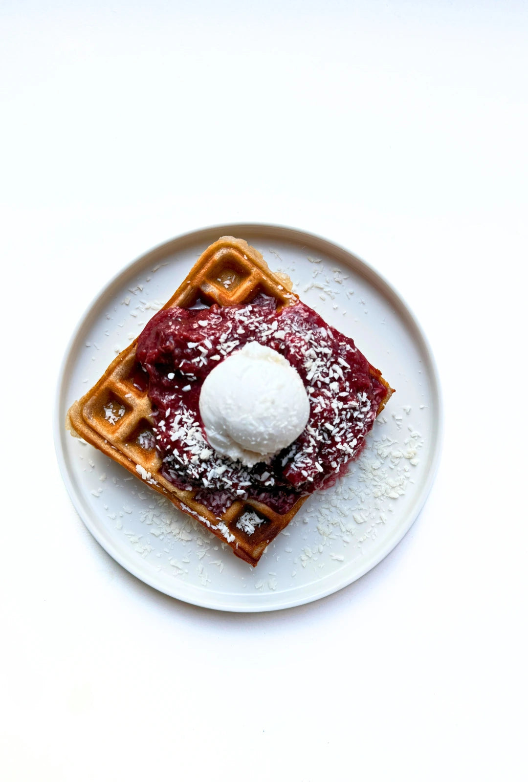 Overhead view of a white plate with a waffle topped with berry compote coconut flakes and a scoop of ice cream