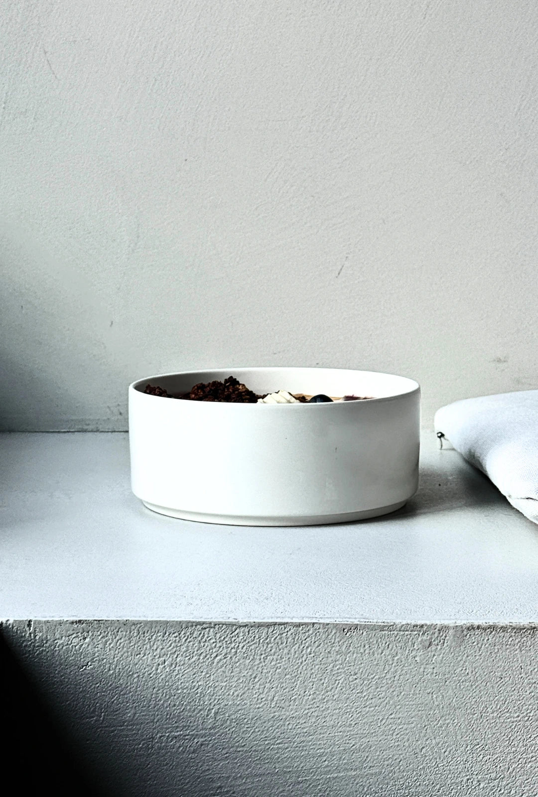 Side view of a white bowl on a pale surface filled with a purple berry smoothie topped with granola