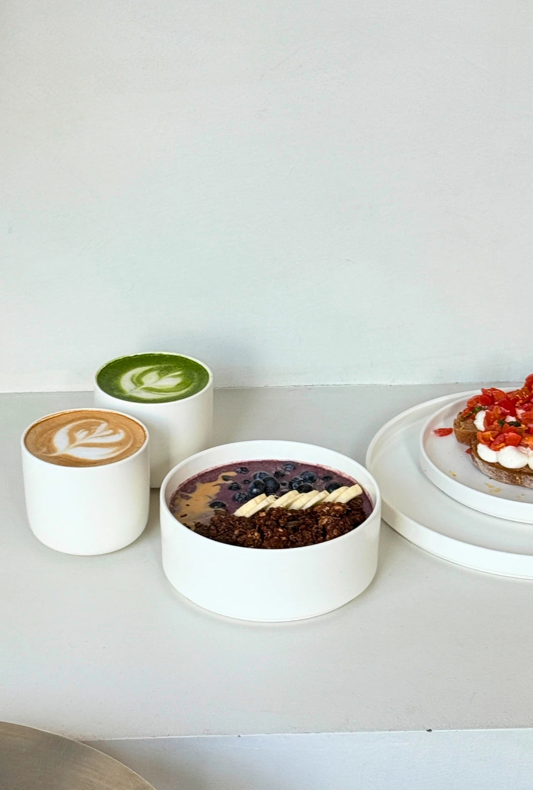 Three white vessels on a pale surface two mugs of hot drinks and a granola berry smoothie bowl beside a pastry on a plate