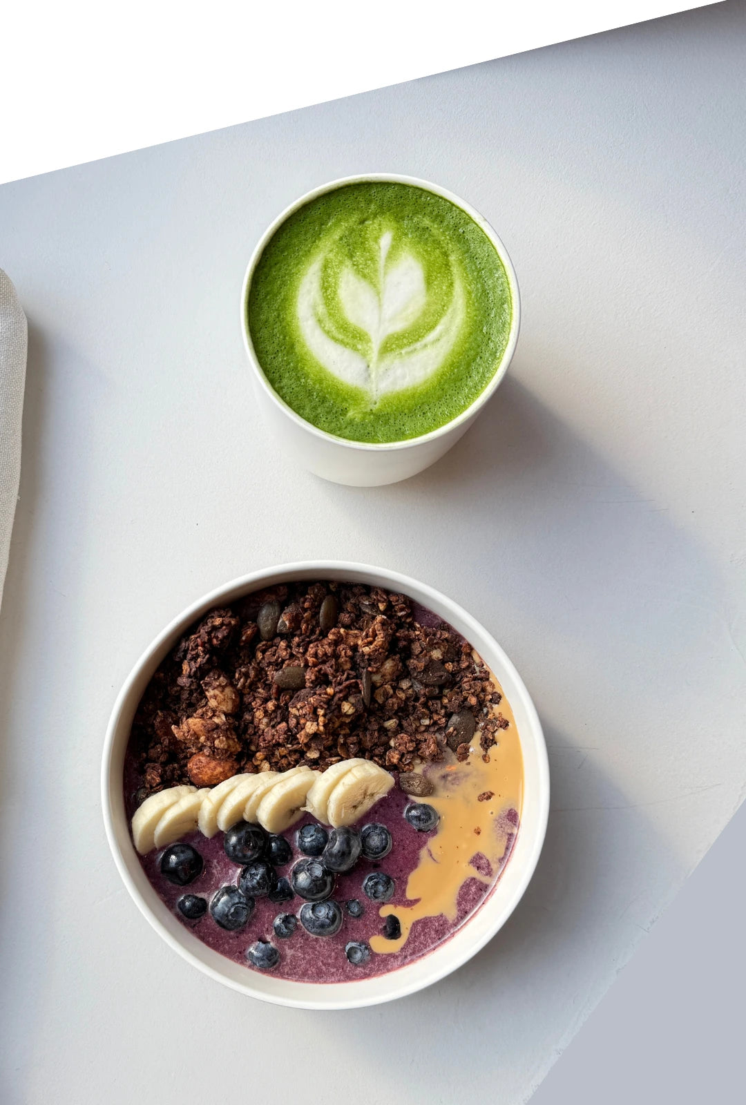 Overhead view of two white bowls on a pale surface a green matcha latte and a berry granola bowl with banana slices and blueberries