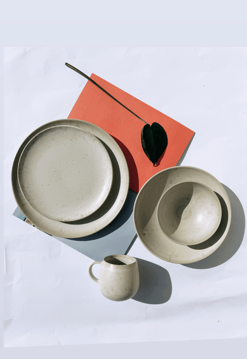 Beige dessert plates with nested bowls and a small mug arranged on a white surface with an orange card and green leaf