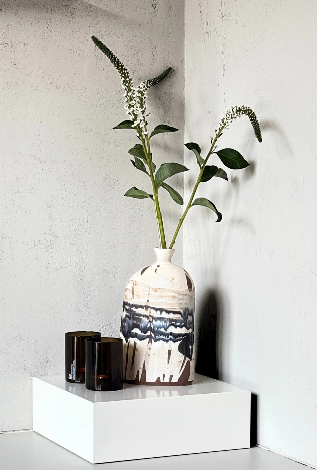 Boa Tarde Vase with tall green stems and white blossoms in a white ceramic vase featuring blue glaze at the bottom on a white pedestal.