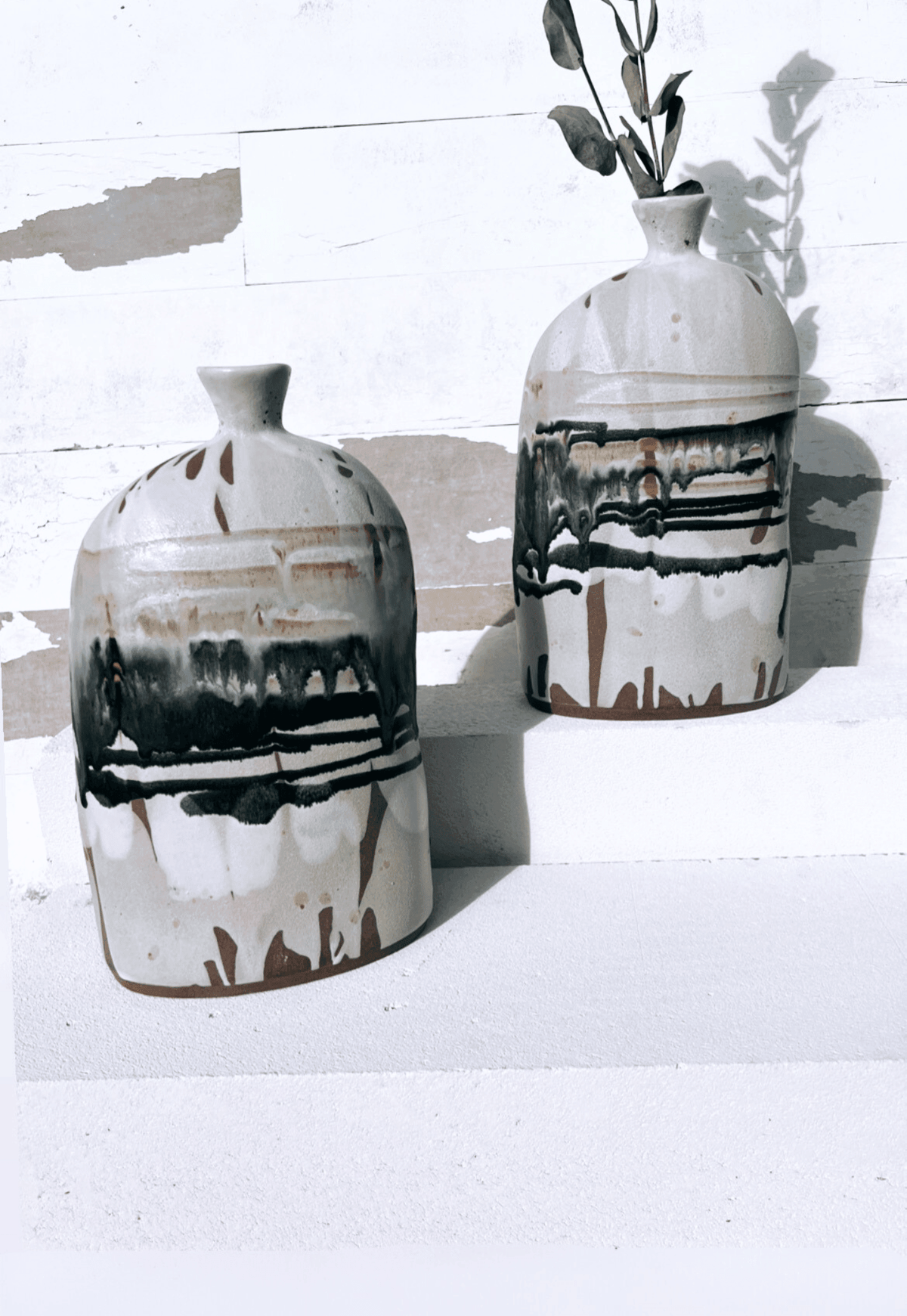 Two Boa Tarde vases with beige glaze and bold drips on a white surface, taller vase showing leafy stems.