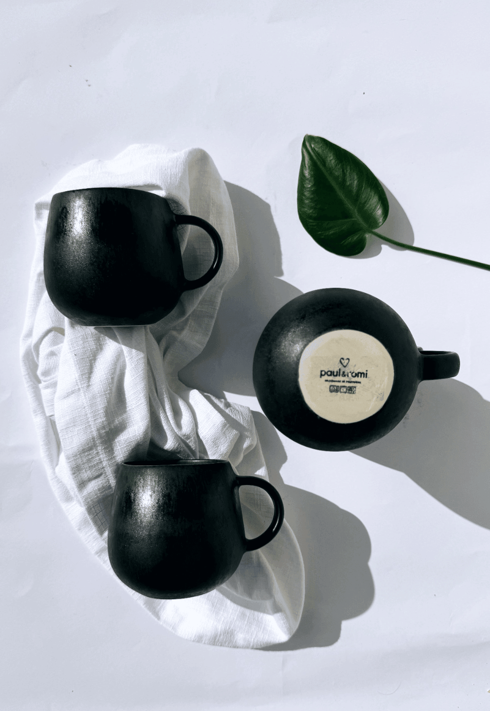 Flat lay of three black ceramic mugs on a white surface with a white cloth and a green leaf, one mug turned over to show its label.