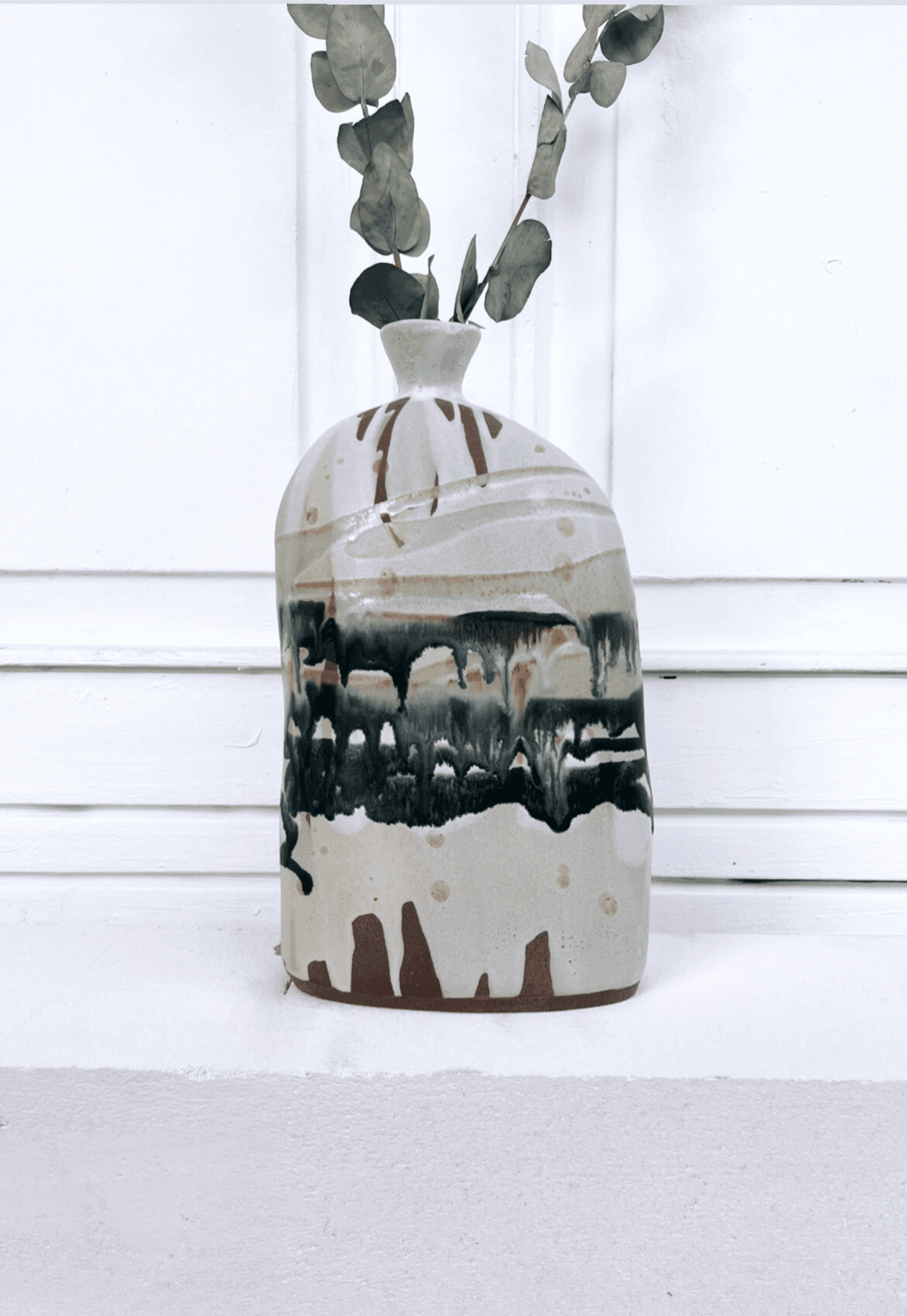 Boa Tarde Vase with pale glaze and brown drip pattern, eucalyptus stems in the narrow neck against a white paneled wall.