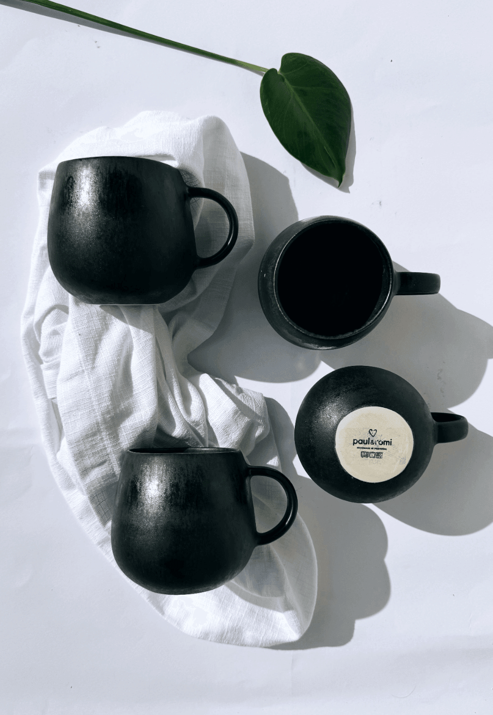Three matte black Alfama Tasse ceramic mugs arranged on a white cloth with a green leaf