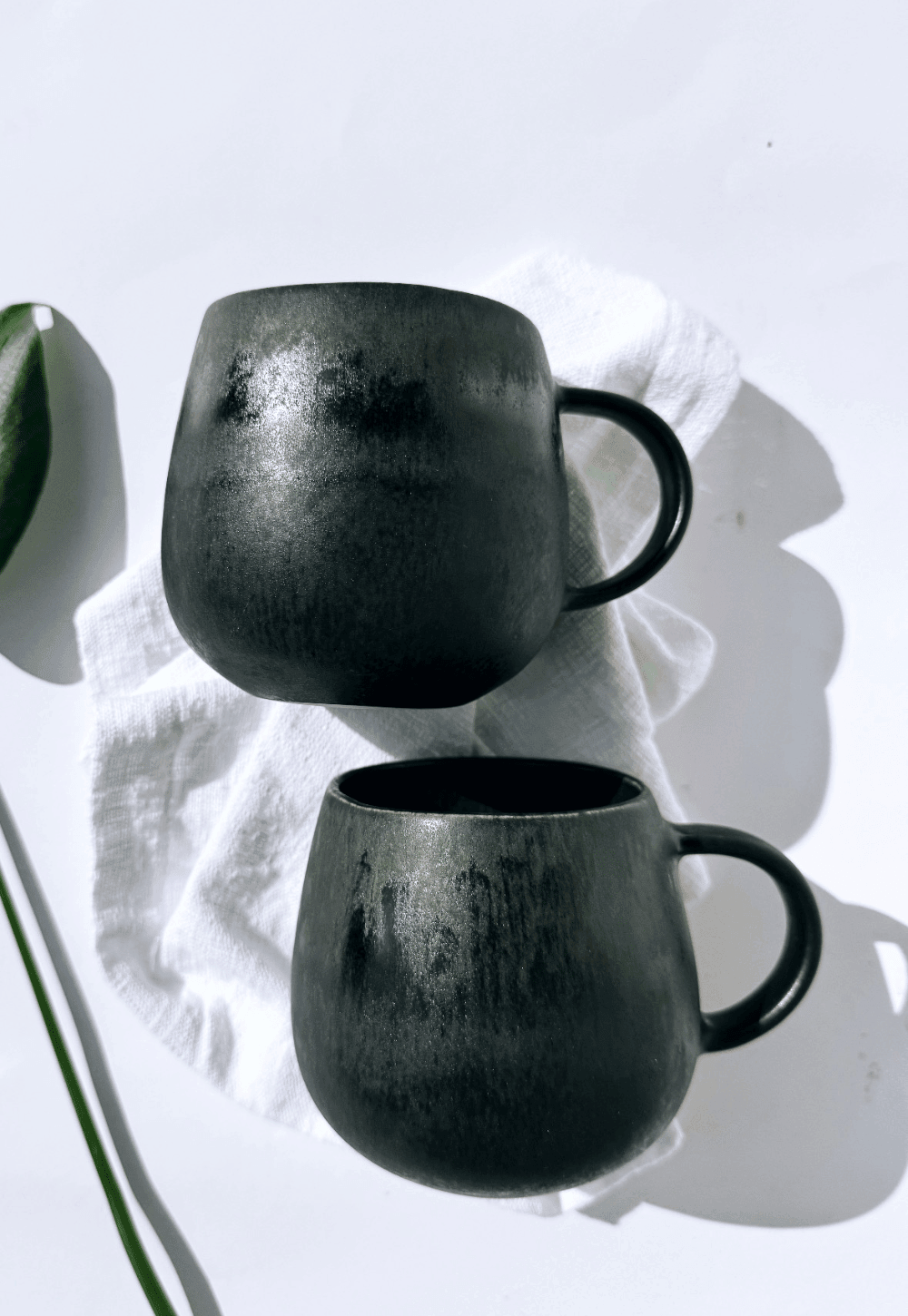 Two matte black ceramic mugs stacked on a white surface with a white cloth and a green plant stem nearby.