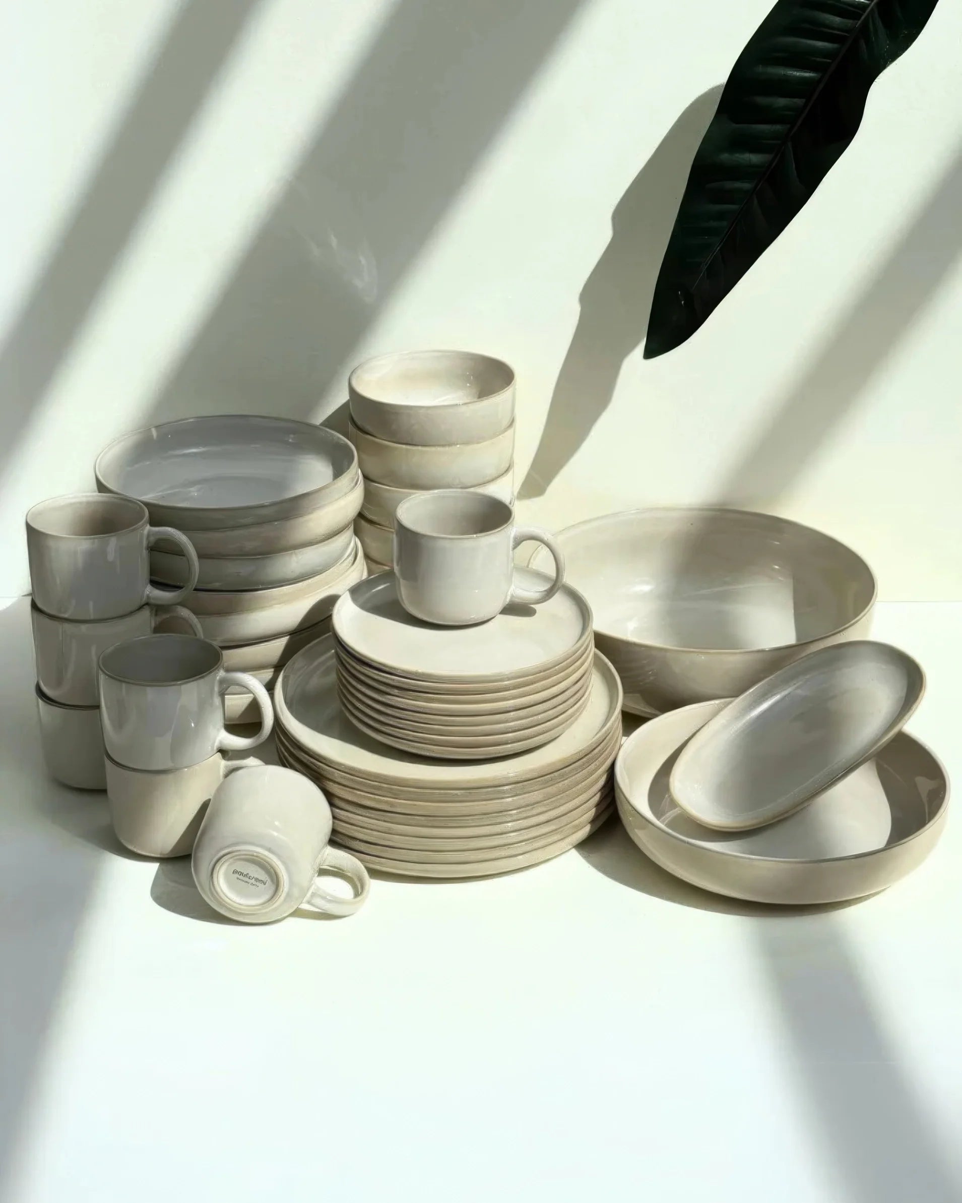 Stack of cream ceramic plates bowls and mugs with a dark green leaf shadow across a pale wall