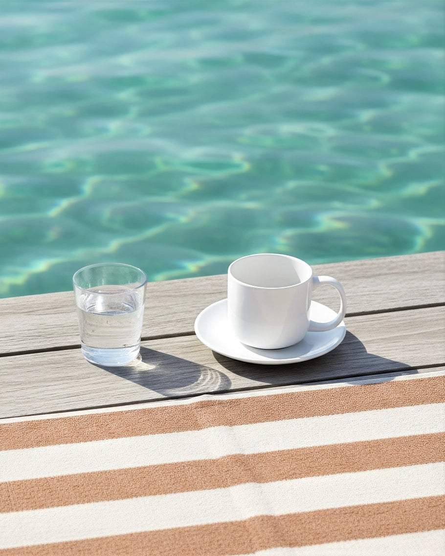 White ceramic mug on saucer beside a glass of water on a sunlit poolside wooden deck.
