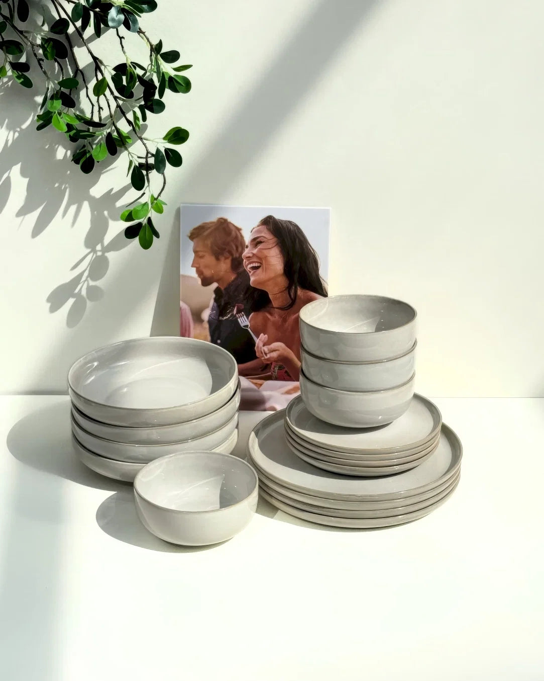 Stacked Scandic Sails bowls and plates on a white surface with a framed photo and a leafy plant in the background.